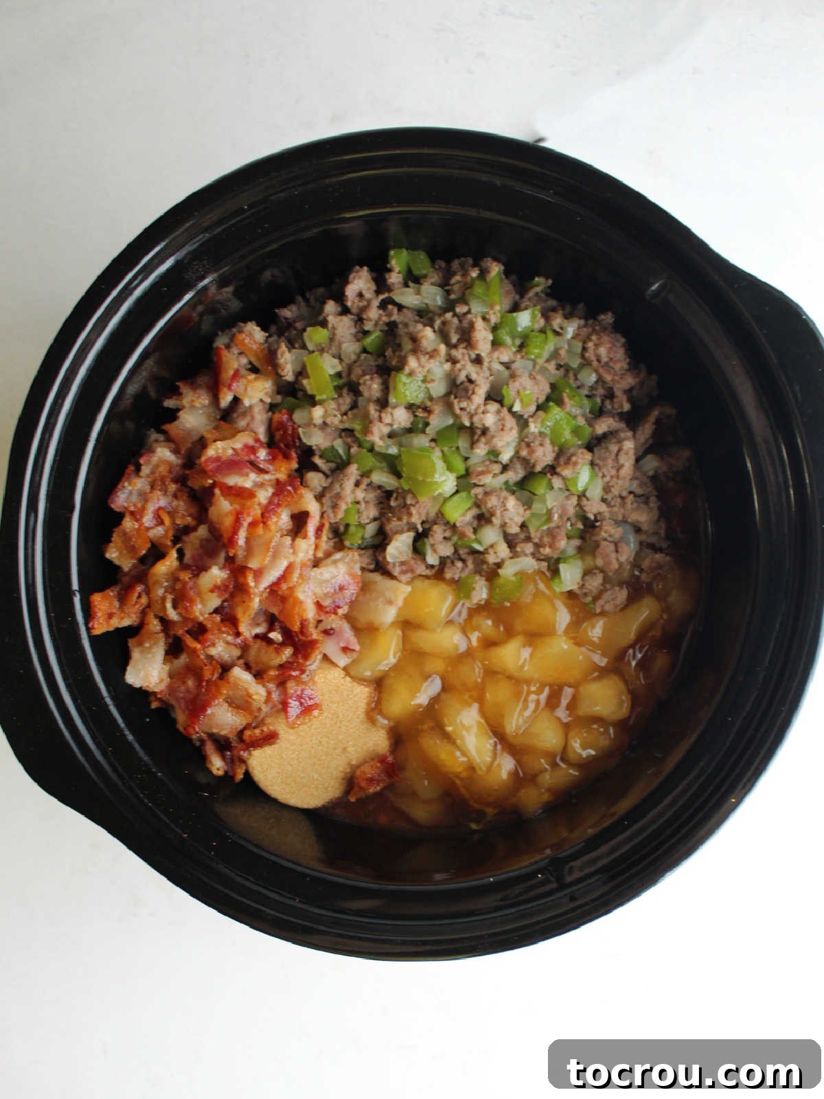 Ultimate Slow-Cooked Baked Beans 5 The process of adding crumbled bacon, cooked sausage, diced onions, and green peppers into a slow cooker, combining savory ingredients for the baked beans.