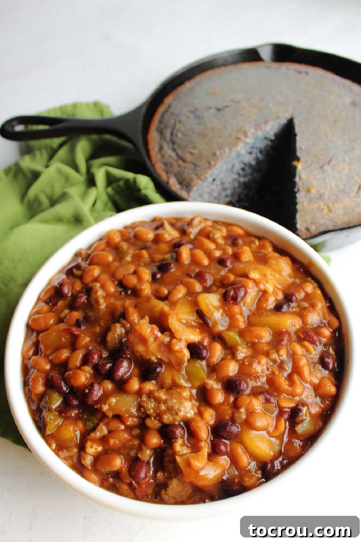 Ultimate Slow-Cooked Baked Beans 7 A rustic bowl overflowing with loaded baked beans, accompanied by a cast iron skillet holding a freshly baked blue cornbread, creating a comforting meal scene.