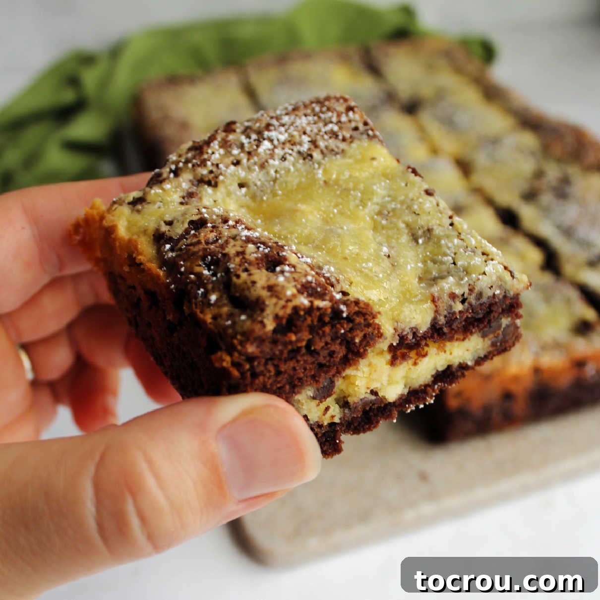 Hand holding a delightful slice of Chocolate Chip Gooey Butter Cake, showcasing its rich chocolate base and creamy, chocolate-studded filling.