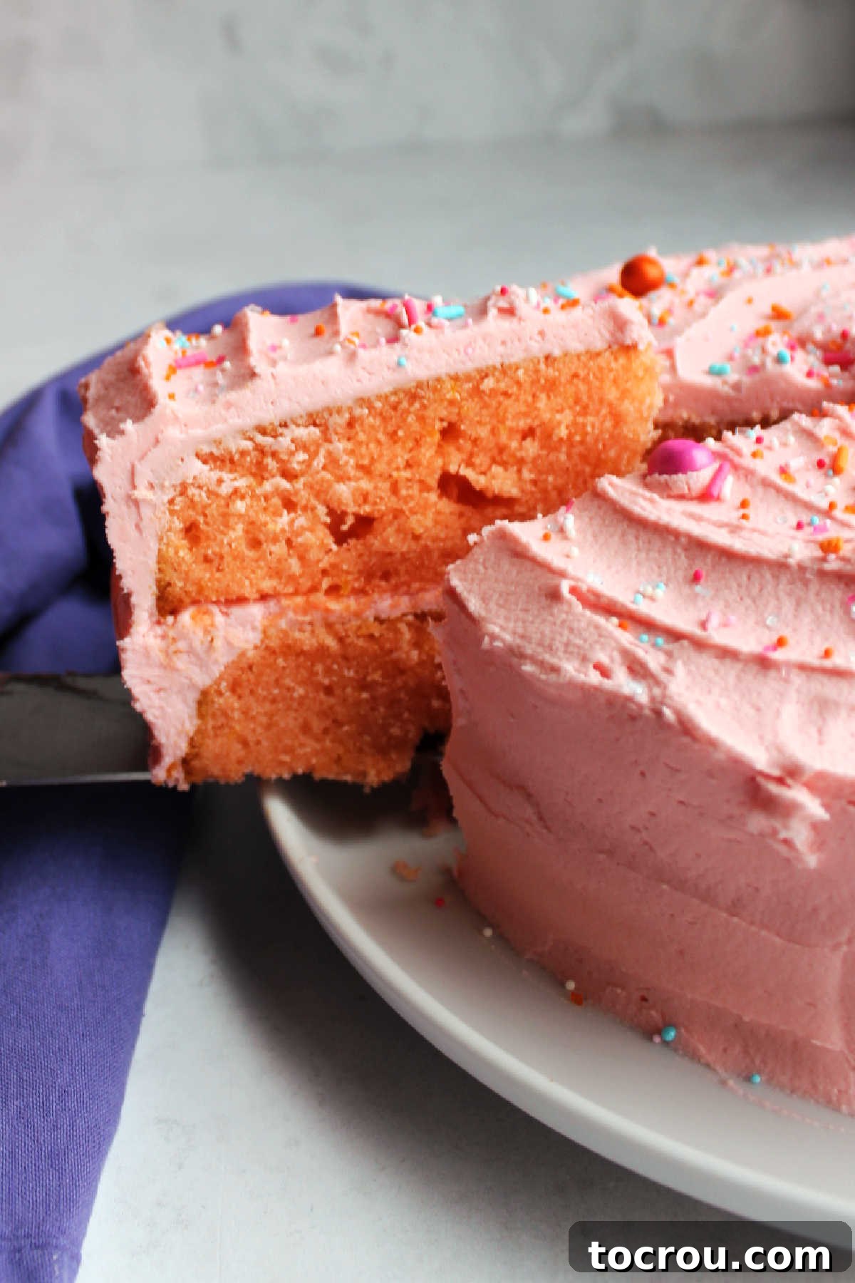 Pulling first slice of pink velvet cake out of the whole cake showing two pink layers of cake and lots of pink buttercream frosting.