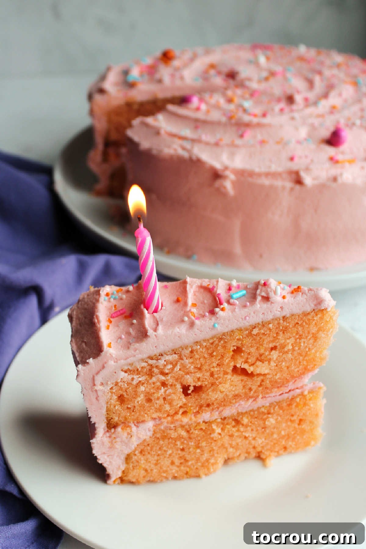 Slice of layered pink velvet cake on plate with lit birthday candle in it. 