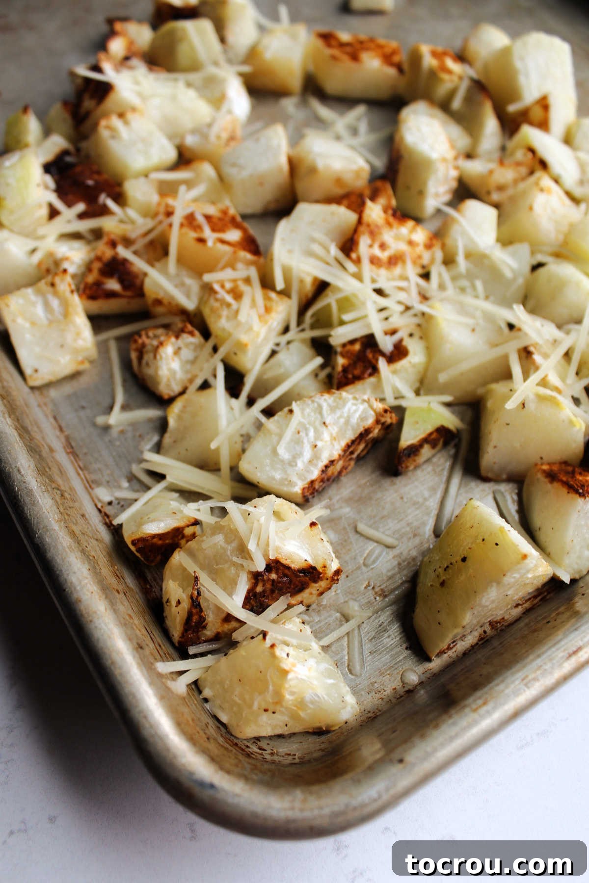 Roasted kohlrabi chunks spread on a pan, lightly sprinkled with grated Parmesan cheese, ready for the final touch in the oven.