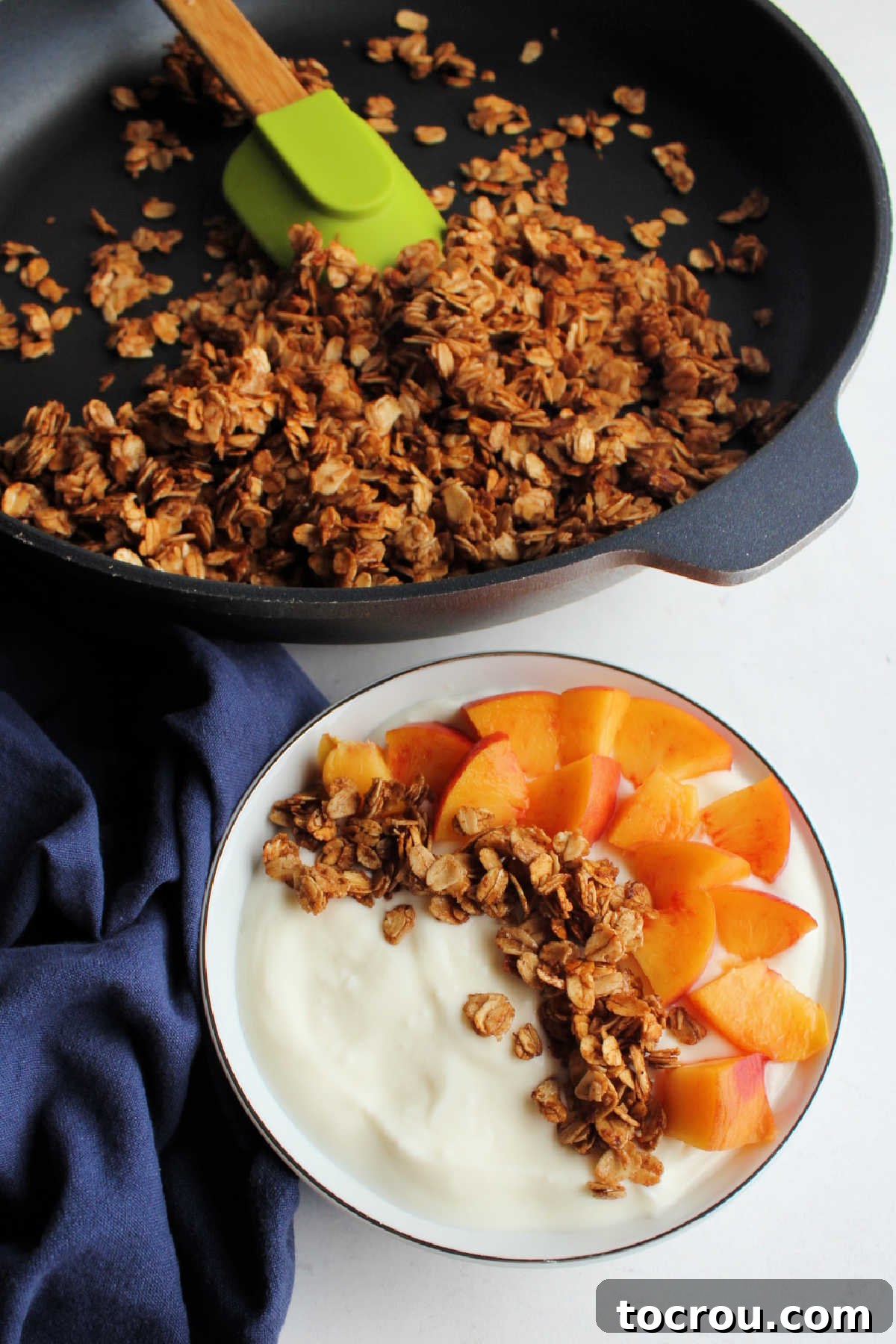 Bowl of yogurt with granola and peaches in front of skillet of remaining granola.