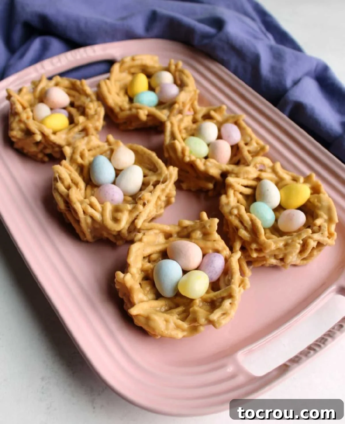 Delicious No-Bake Peanut Butter Chow Mein Nests for Spring Pink platter filled with vibrant peanut butter and marshmallow chow mein nests, each nestled with colorful candy eggs, perfect for a festive spring or Easter display.