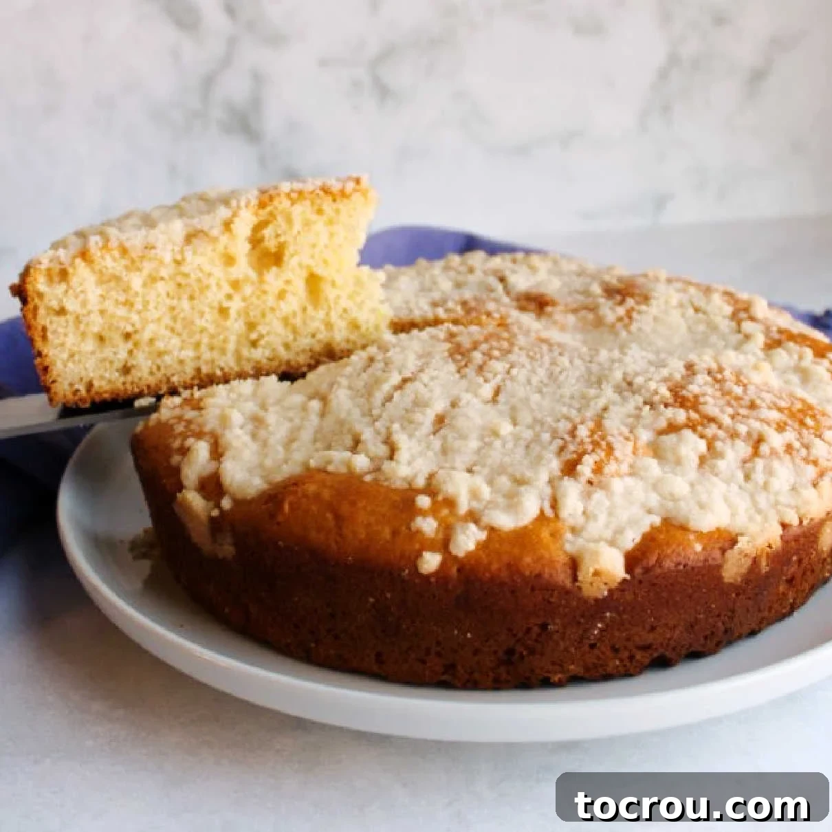 Enjoy a slice of traditional Polish Placek, a sweet bread with buttery crumb topping perfect for any occasion. First slice of crumb-topped Polish Placek being lifted from a round, cake-like loaf, showcasing its soft interior and golden streusel.