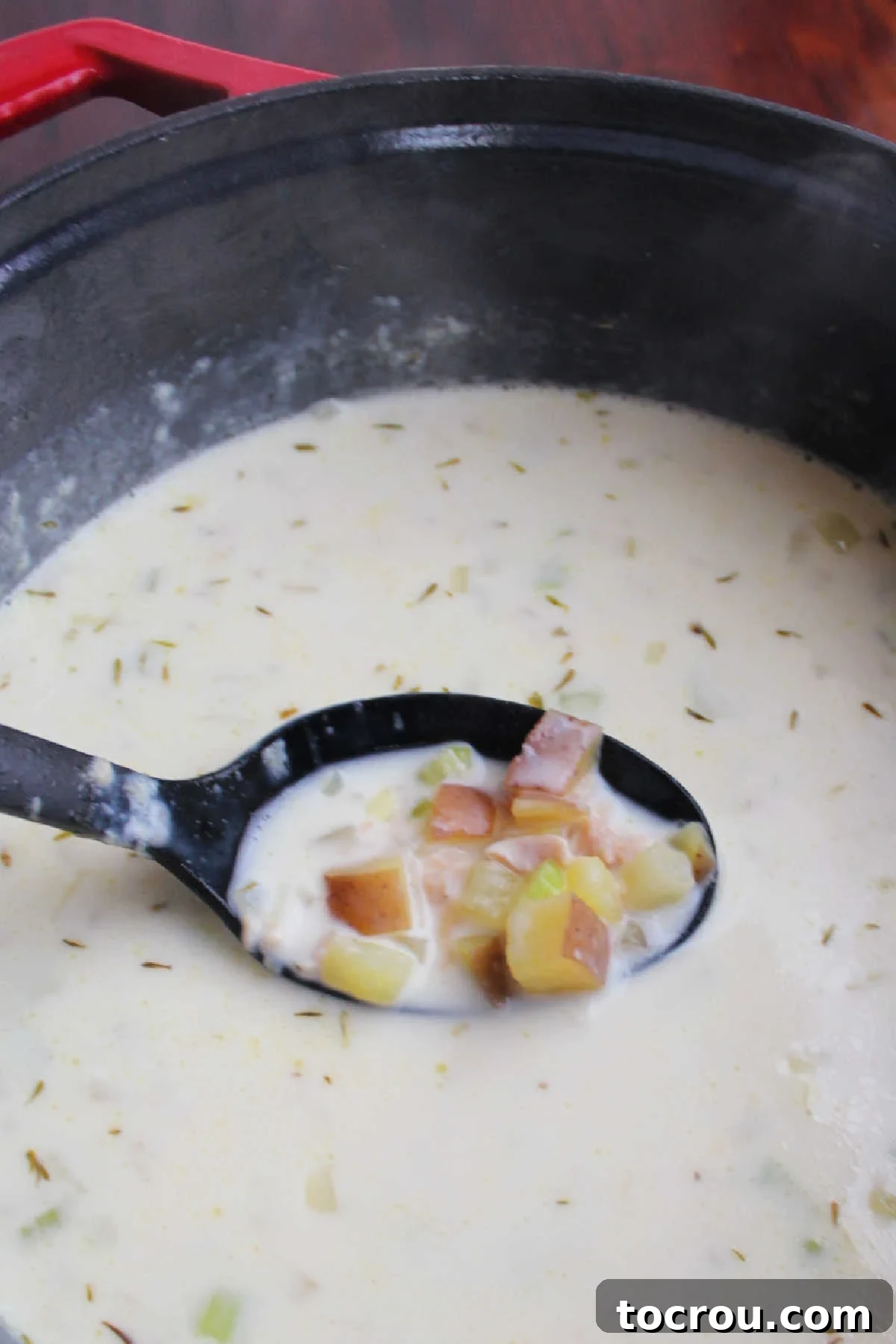 Serving spoon lifting out chunks of potatoes, celery, and onions out of an enameled dutch oven full of new England clam chowder.