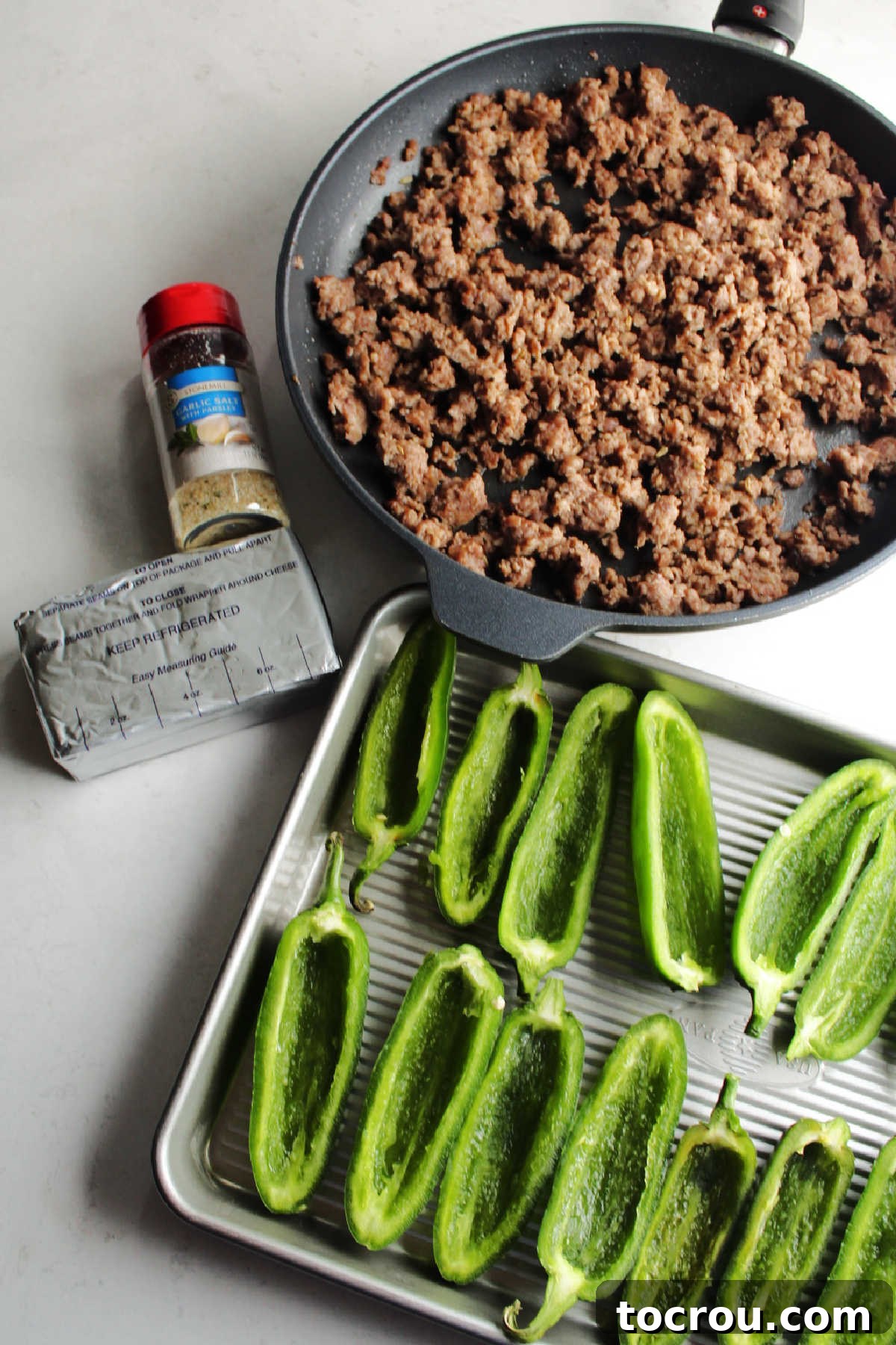 Ingredients laid out on a counter: browned sausage crumbles, a block of cream cheese, a small bowl of garlic powder, and halved, cleaned jalapeno peppers, all ready for assembly.