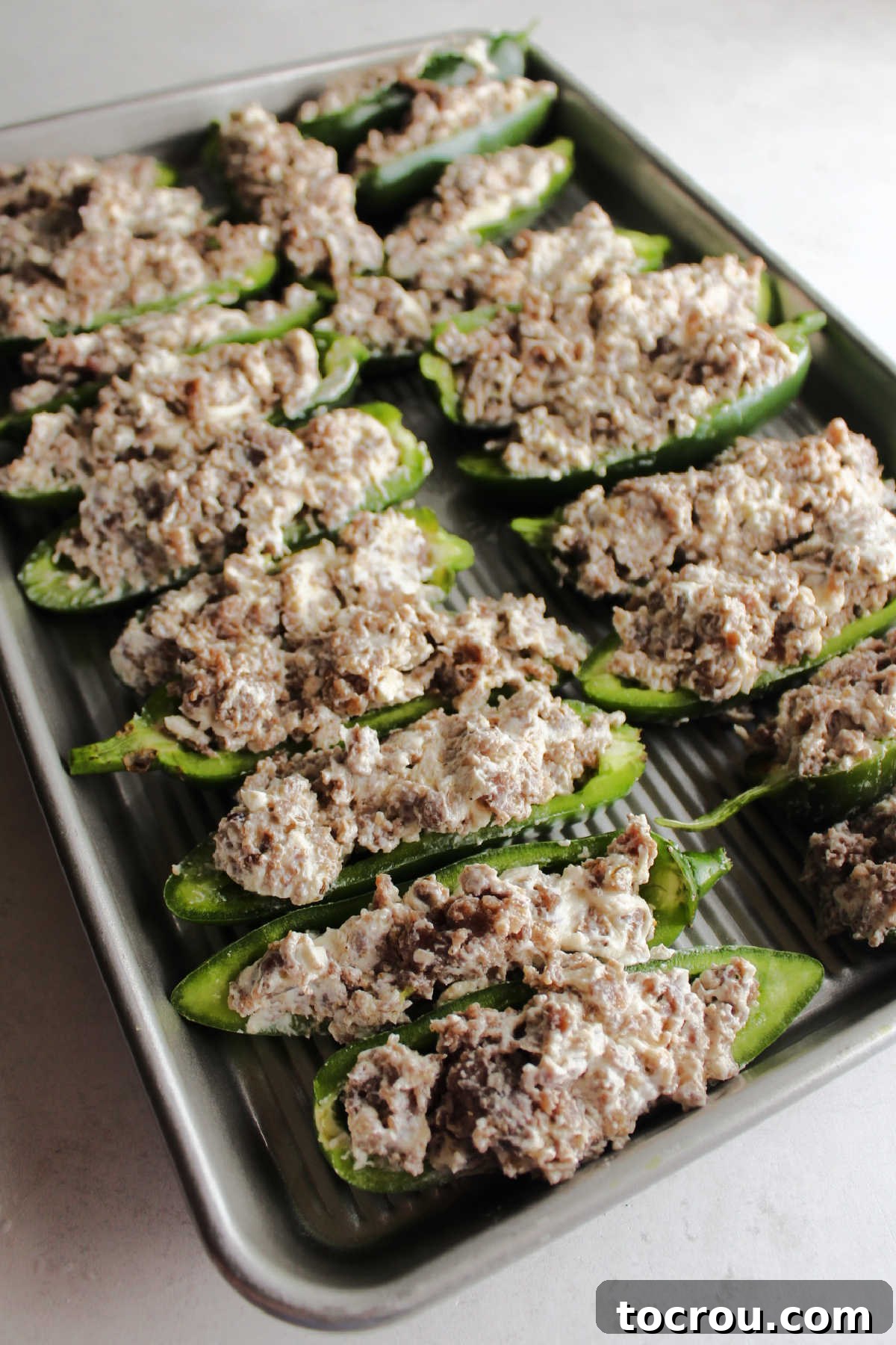 A baking tray filled with numerous halved jalapeno peppers, generously stuffed with the sausage and cream cheese mixture, ready to be baked in the oven.
