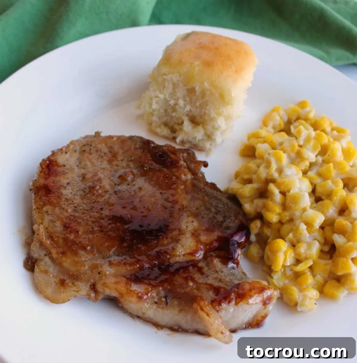 Tender and Flavorful Braised Pork Chops with Savory Sauce Close up of a perfectly braised pork chop with a glossy brown glaze, served alongside fresh corn and a golden Hawaiian roll on a dinner plate.