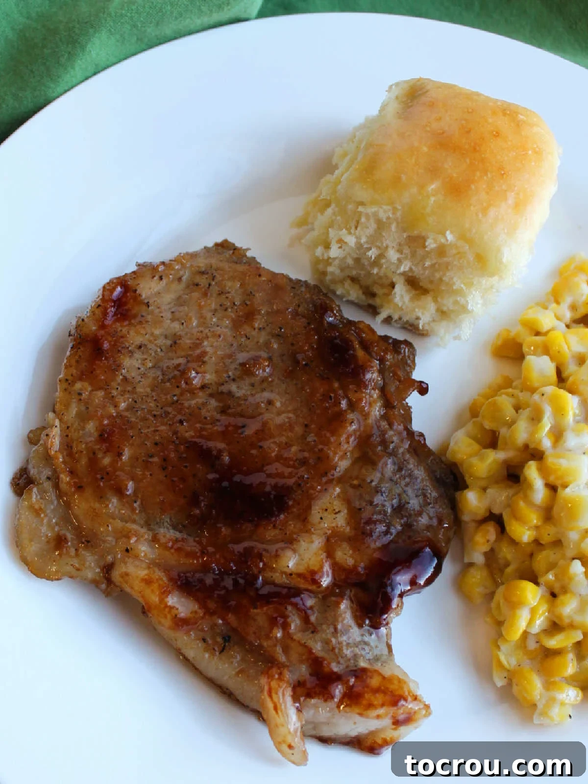 Braised Pork Chop with Homestyle Sides Dinner plate showcasing a large, beautifully glazed braised pork chop, served with creamy cheesy corn and a fluffy homemade pineapple Hawaiian dinner roll.