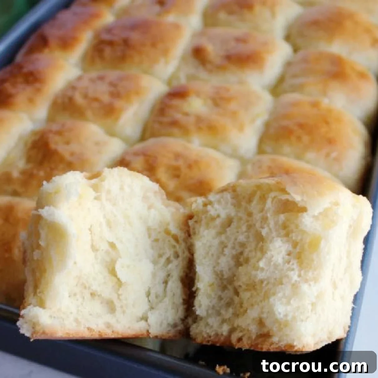 Close-up of the soft, golden interior of a homemade pineapple Hawaiian roll, with a pan of rolls in the background.