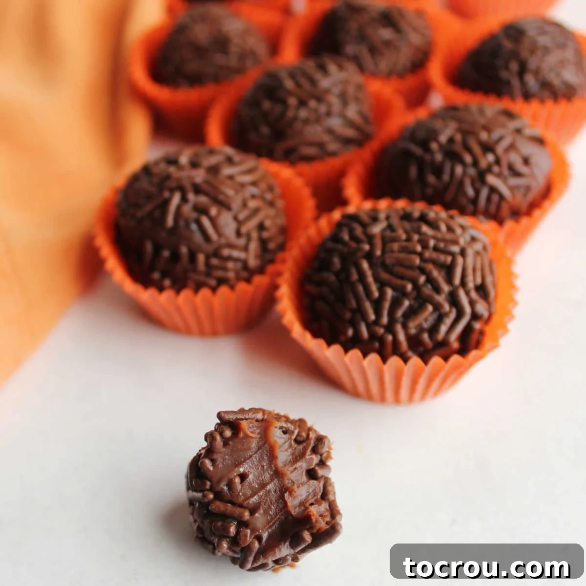 Brigadeiros are a delicious chocolate candy from Brazil. They are easy to make and require just a few pantry staples as ingredients. Add a hint of orange to make them an extra special treat! Close-up of a chocolate orange brigadeiro, cut open to reveal its soft, creamy condensed milk center. More sprinkle-coated truffles are blurred in the background.