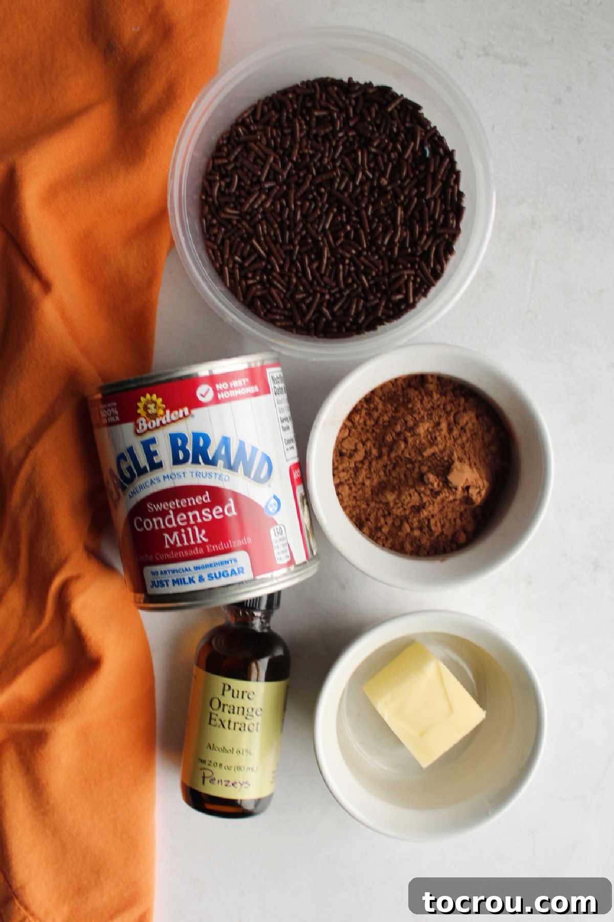 Zesty Chocolate Brigadeiro 3 Key ingredients laid out: a can of condensed milk, a bowl of cocoa powder, a stick of butter, a bottle of orange extract, and a dish of chocolate sprinkles, ready for brigadeiro making.