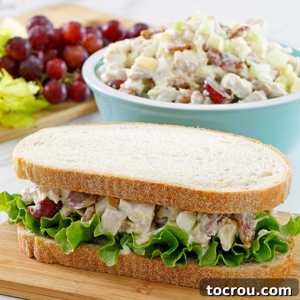 A vibrant pineapple chicken salad sandwich, artfully arranged with crisp lettuce, sits ready to be enjoyed. In the background, a rustic bowl overflows with more of the delicious fruited chicken salad, showcasing its appealing blend of colors and textures.