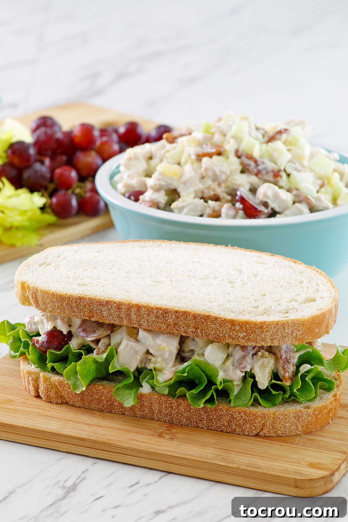 A hearty sandwich crafted with thick bread, fresh lettuce, and our signature chicken salad featuring pineapple and grapes. It's elegantly presented in front of a full bowl of the chicken salad and a lush bunch of grapes, emphasizing its fresh ingredients.
