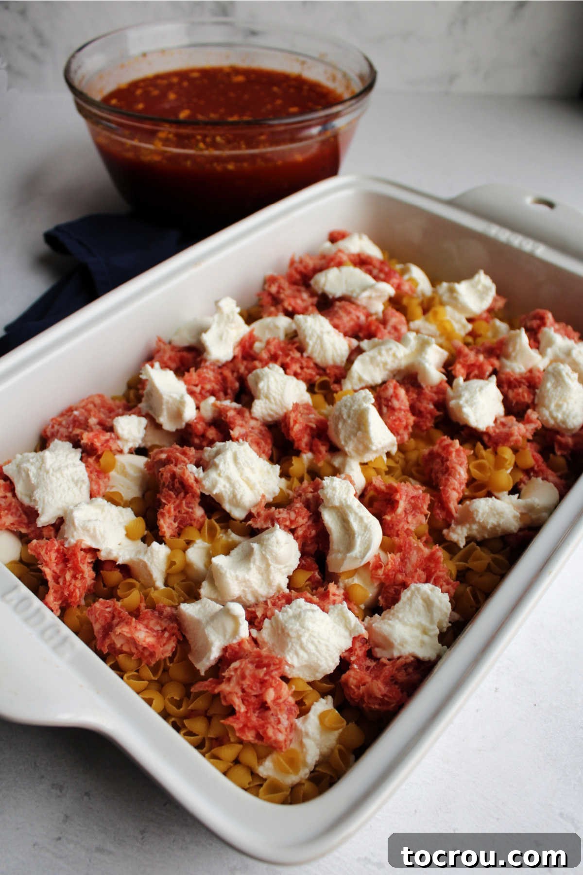 Prepping the no-boil casserole ingredients uncooked shell pasta, raw sausage and chunks of cheese in baking dish with bowl of sauce in background.