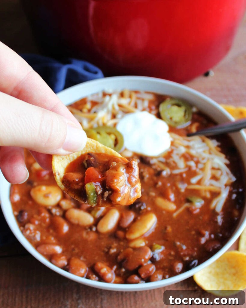 Hand holding a corn chip that has been scooped into the chili with some chili inside, ready to eat. 