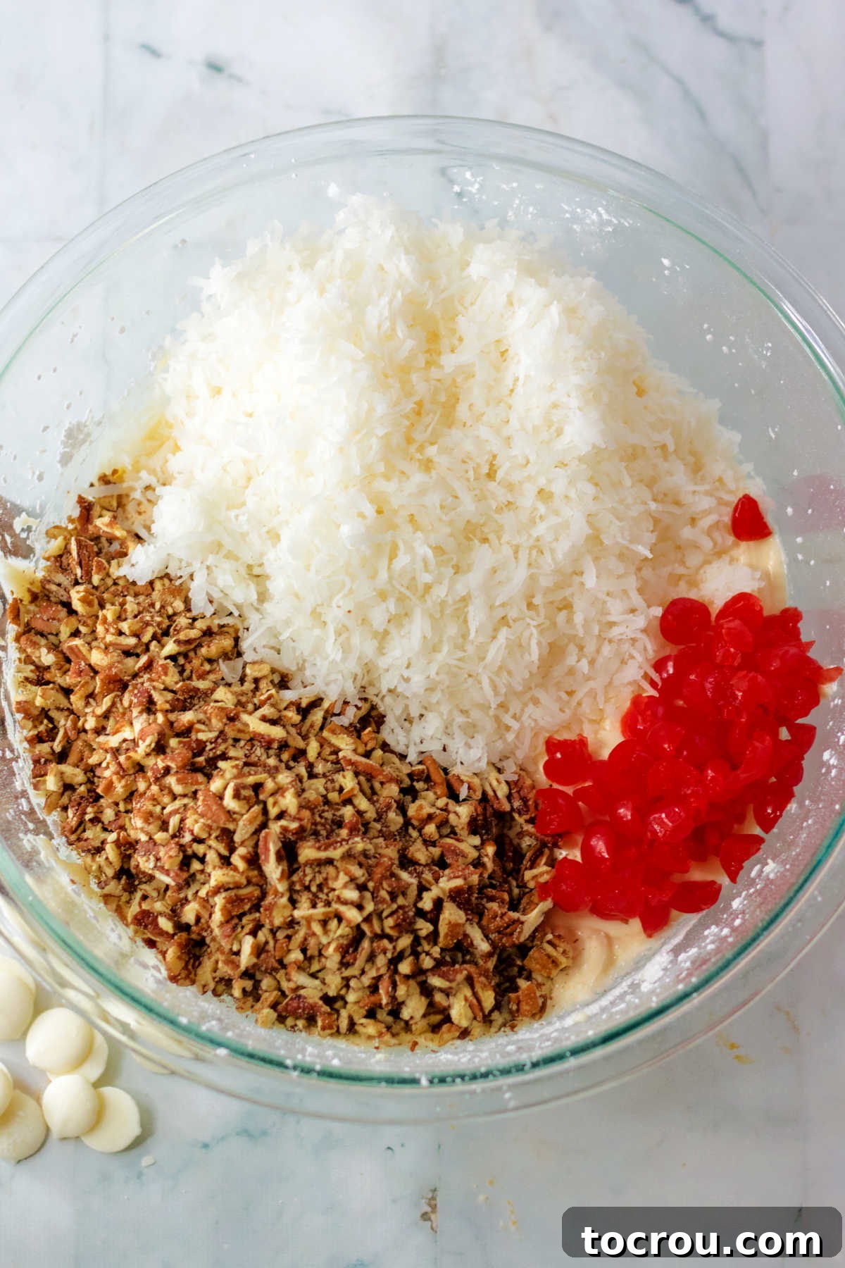 Adding shredded coconut, finely chopped maraschino cherries, and pecan bits to the creamy butter and powdered sugar mixture in a glass bowl.