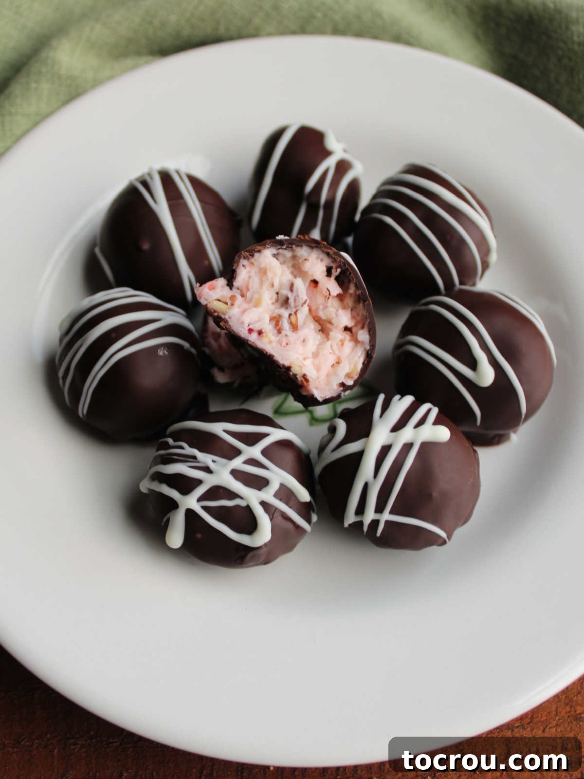 A plate of chocolate-dipped Martha Washington candies with white chocolate drizzle, with one candy cut in half nearby to reveal its creamy cherry, coconut, and pecan buttercream center.