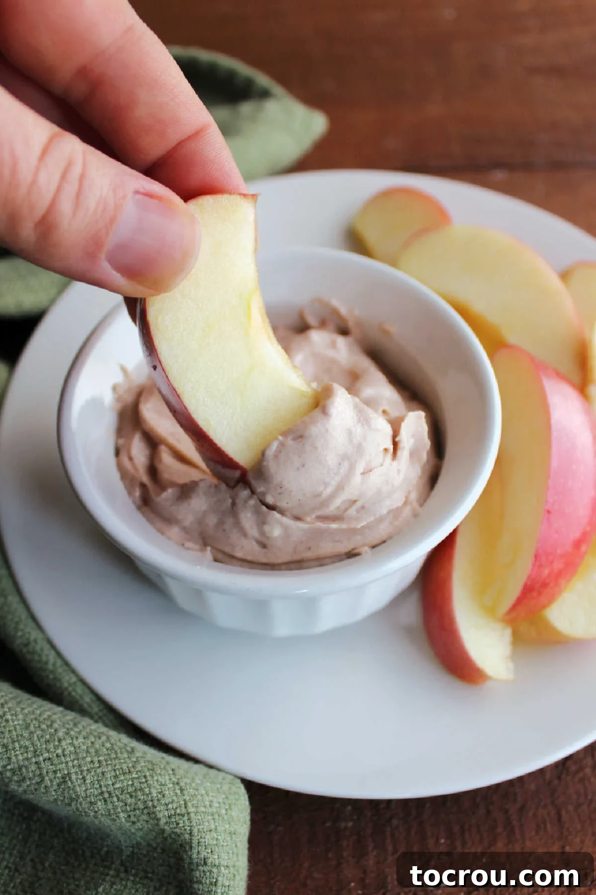 A hand gracefully dipping a slice of fresh apple into a small ramekin of smooth, inviting cream cheese apple butter dip.