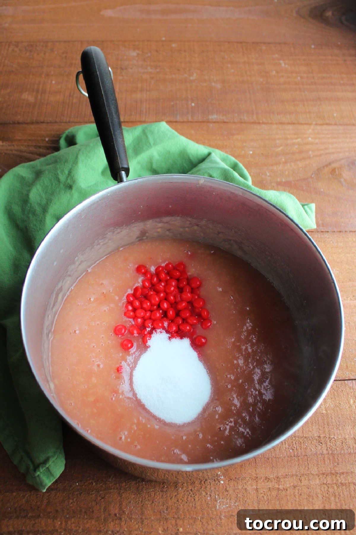 Granulated sugar and vibrant Red Hot candies being added to a saucepan filled with pink applesauce.