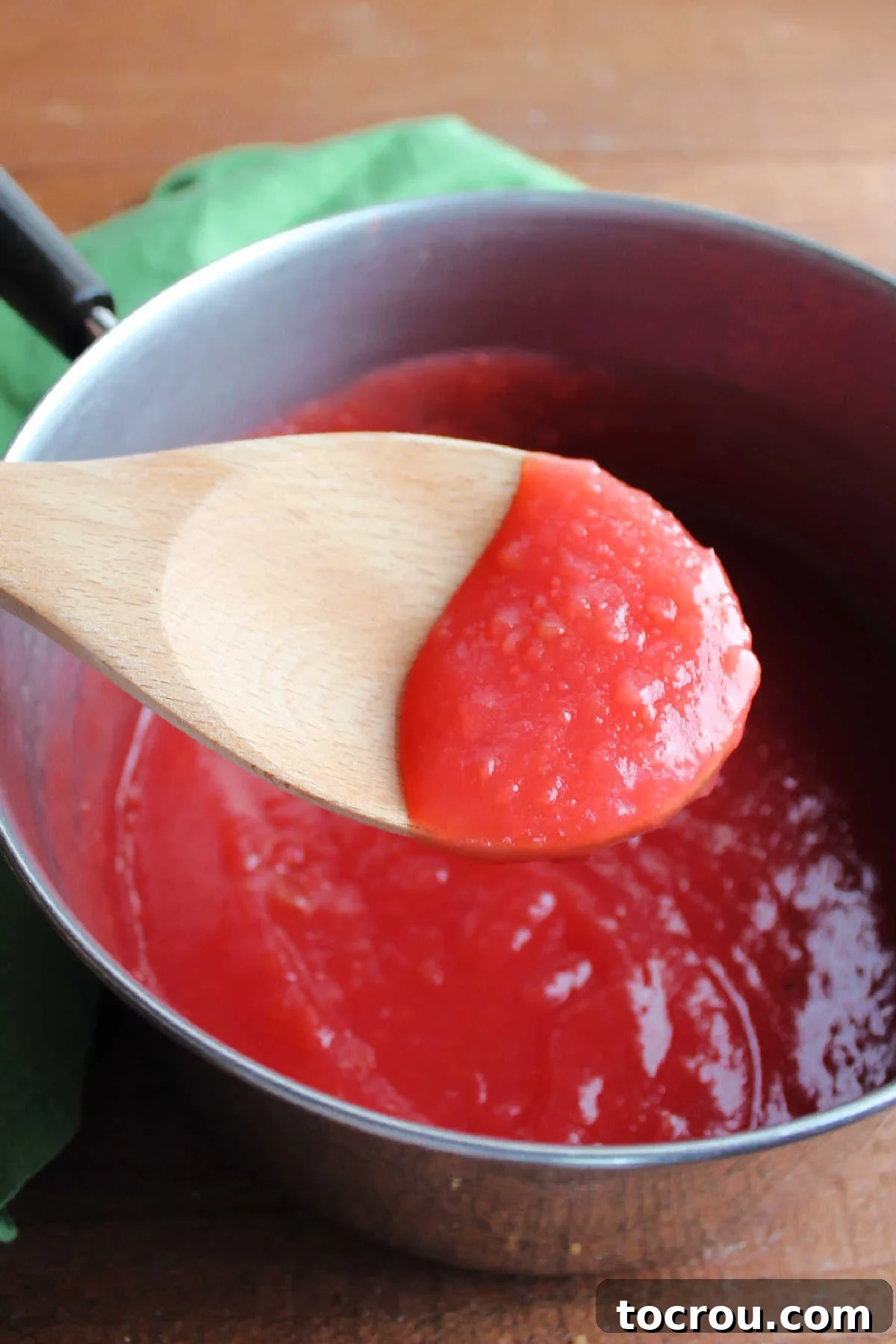 A wooden spoon holding a spoonful of freshly made red hot applesauce, hovering over a saucepan of the smooth, sweet, and spicy sauce.
