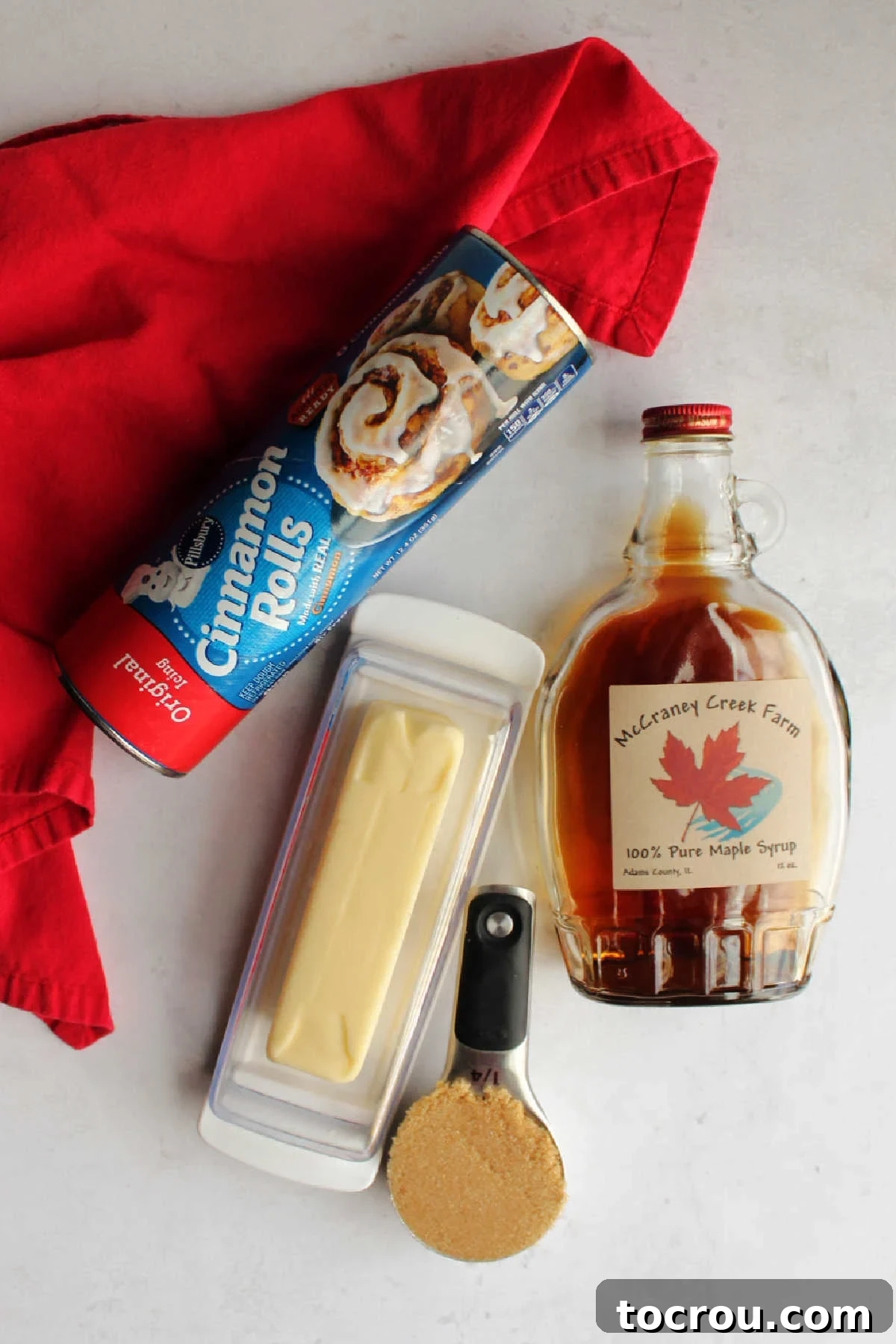 The four essential ingredients for easy sticky buns displayed on a kitchen counter: a tube of cinnamon rolls, a stick of butter, a bowl of brown sugar, and a bottle of pure maple syrup.