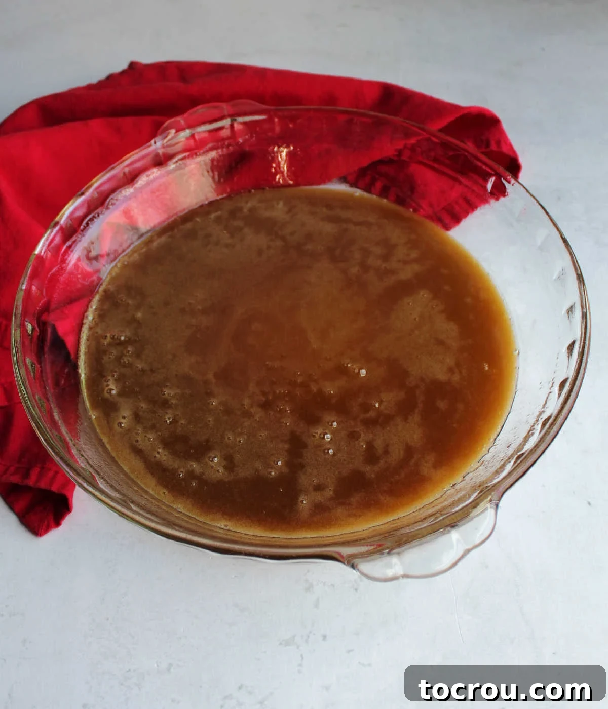Close-up of the smooth, amber-colored maple, brown sugar, and butter sauce poured into the bottom of a clear glass pie dish, ready for the cinnamon rolls.