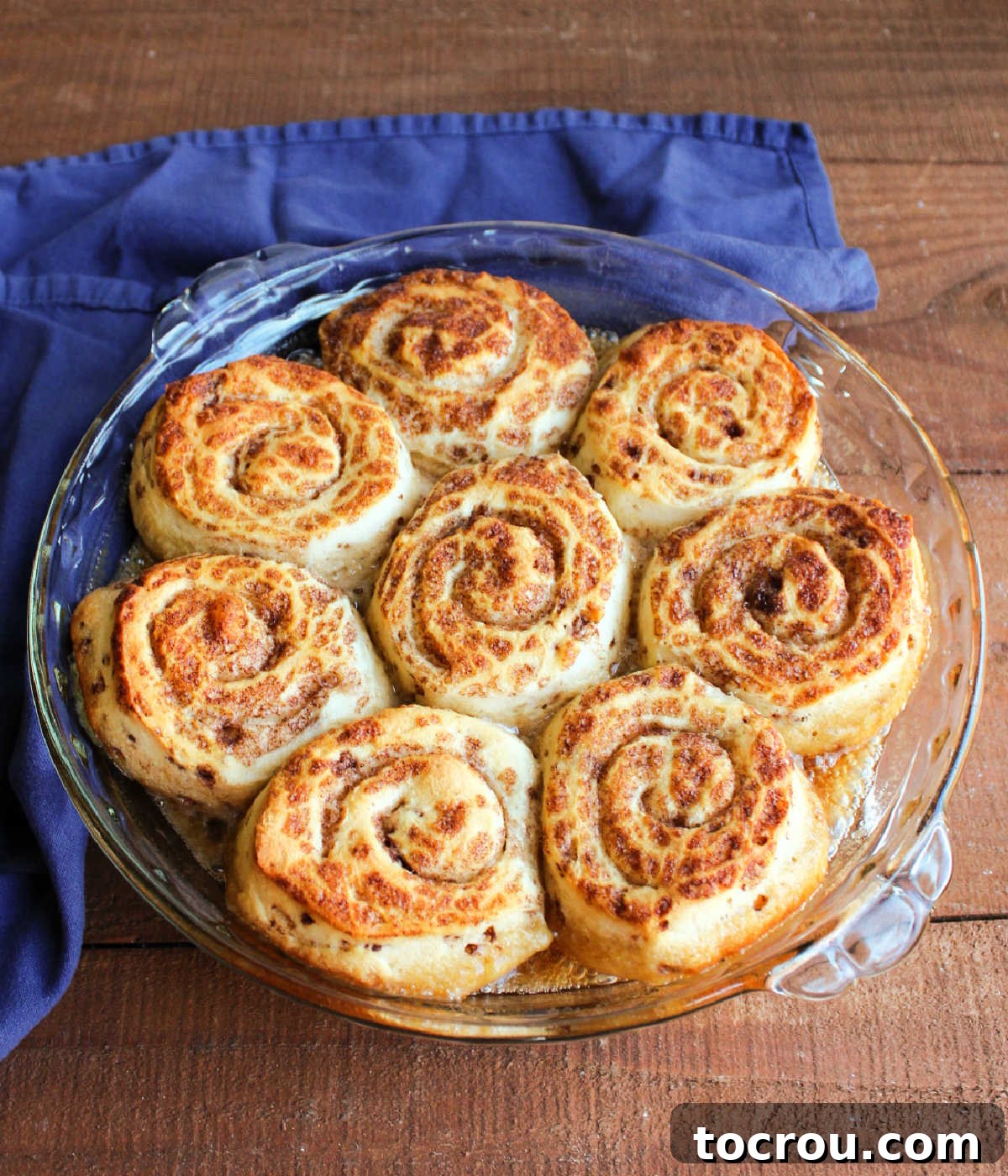 A freshly baked batch of maple sticky buns glowing golden-brown in a glass pie plate, their surfaces puffed and ready to be inverted, promising a cascade of gooey maple sauce.
