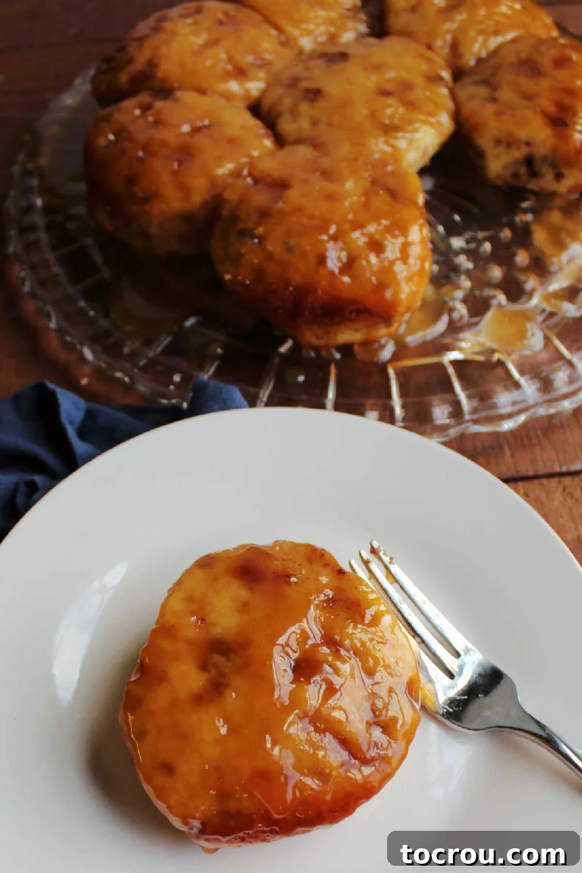 A close-up shot of a single maple sticky bun on a plate, adorned with a generous, thick layer of glistening maple caramel sauce, with the remaining sticky buns blurred in the background.