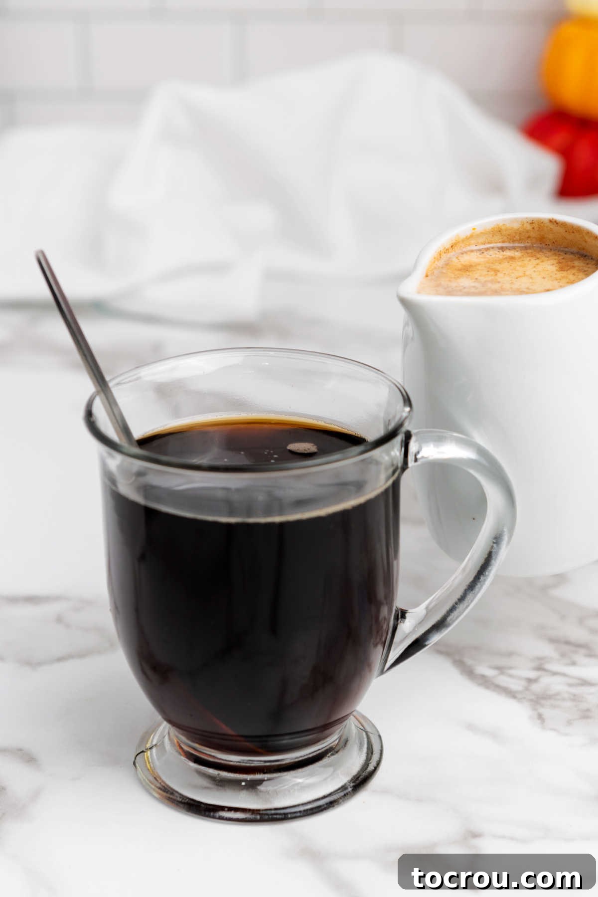 Glass mug filled with coffee, with a pitcher of homemade pumpkin spice creamer ready to be poured in, highlighting the rich liquid texture. 