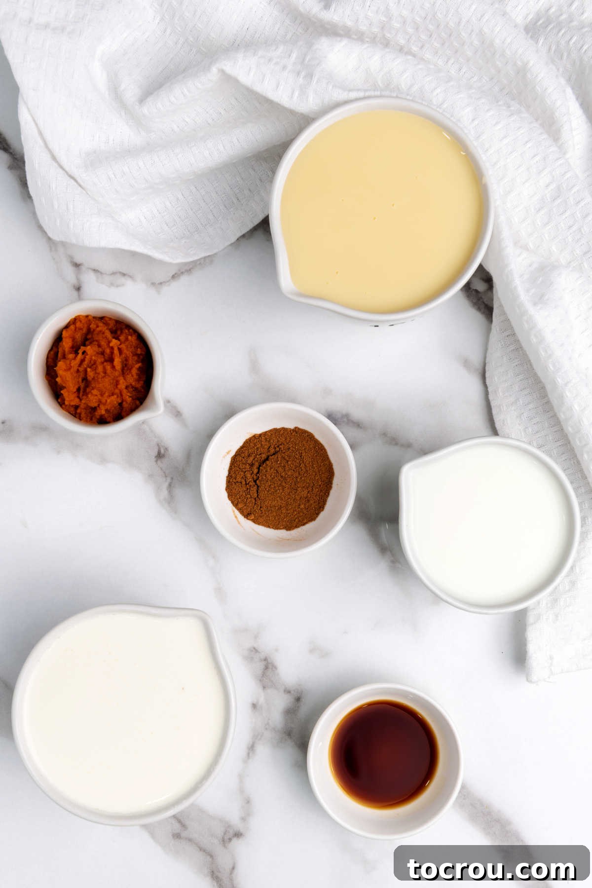 Individual ingredients like a can of pumpkin puree, a bottle of vanilla extract, and a jar of pumpkin pie spice, neatly arranged before being mixed into creamer.