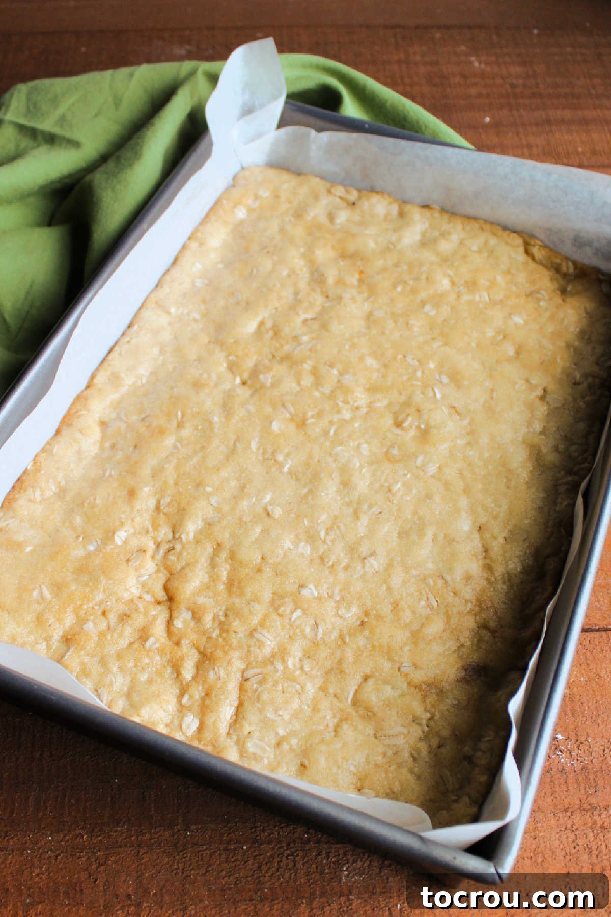 The partially baked oatmeal cookie layer in the pan, a light golden brown and ready for the rich caramel, chocolate, and pecan toppings.