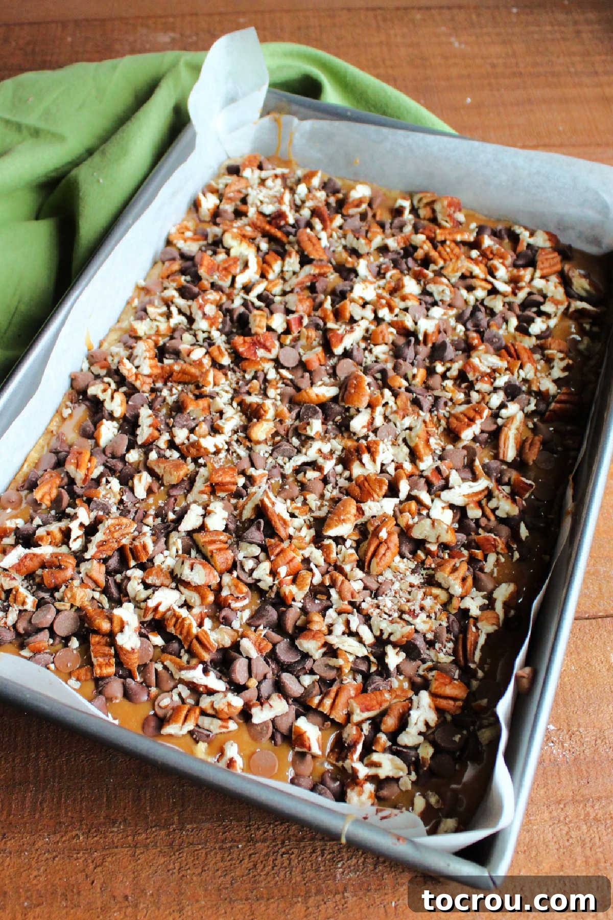 The pre-baked oatmeal cookie layer generously topped with a smooth, melted caramel, followed by a sprinkle of chocolate chips and chopped pecans, forming the delicious middle layer.