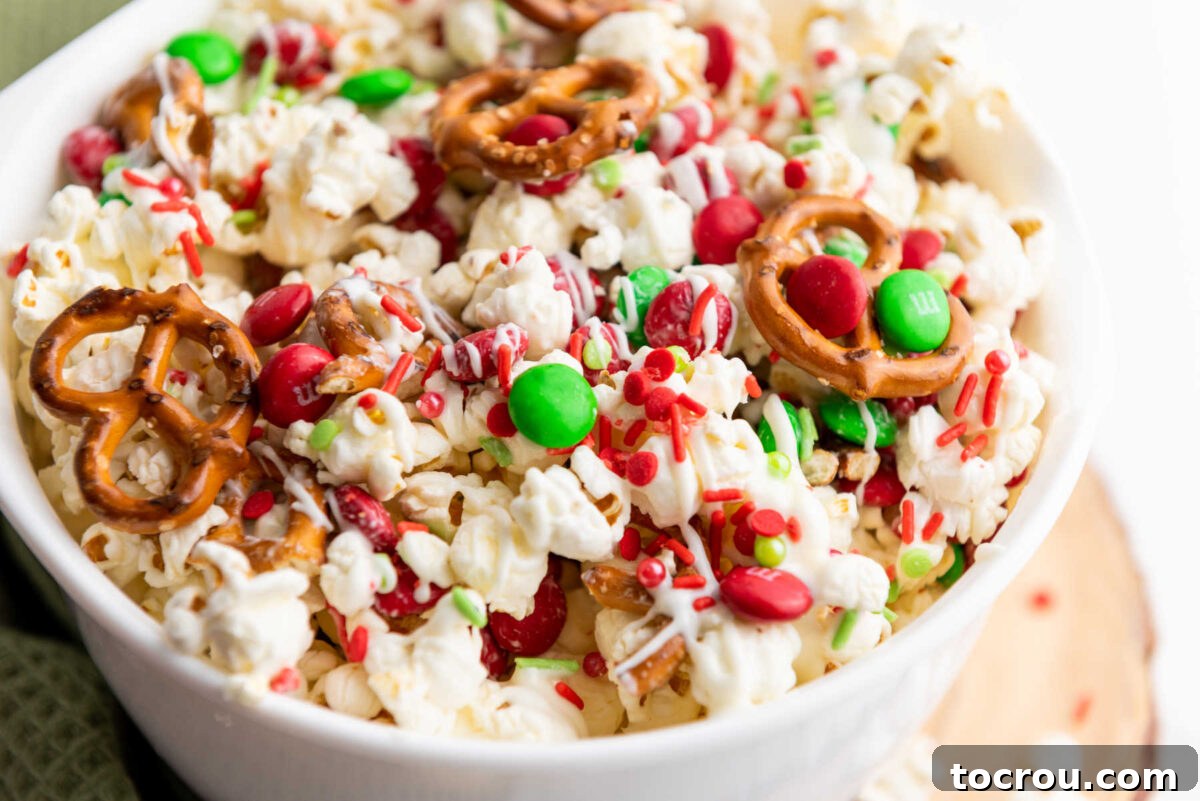 Close up of a bowl of popcorn with pretzels, white chocolate drizzle, and holiday M&Ms and sprinkles, ready to eat. A perfect sweet and salty Christmas popcorn snack mix for holiday movie nights and parties.