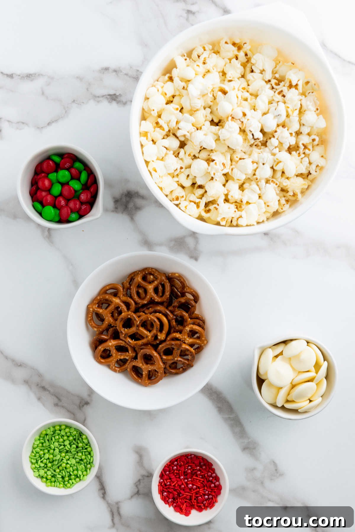 Ingredients: freshly popped popcorn, crunchy pretzels, colorful holiday chocolate candies (like M&Ms), and festive sprinkles laid out on a tray, ready to be transformed into a delicious Christmas snack mix.