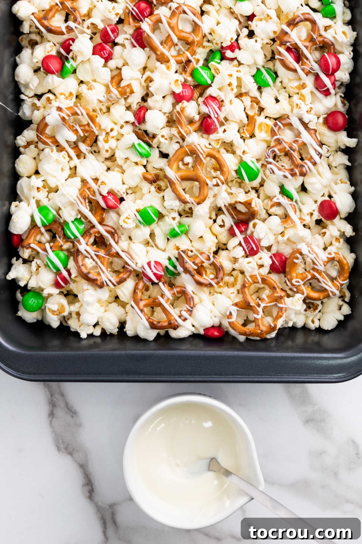 A stream of rich, melted white chocolate being drizzled carefully over a festive mixture of popcorn, pretzels, and holiday M&Ms, preparing for the final touch of sprinkles to create a delightful Christmas snack.