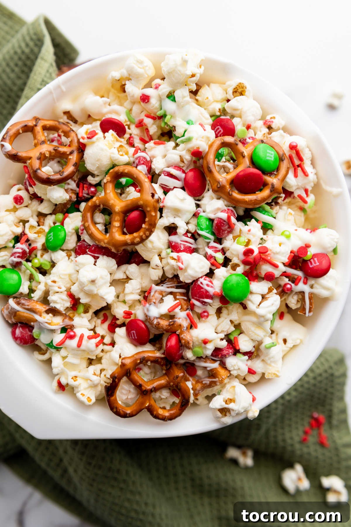 An overhead shot revealing a delightful bowl of Christmas popcorn, highlighting the interplay of fluffy white popcorn, crunchy pretzels, and vibrant red and green holiday M&Ms, all coated with a delicate white chocolate drizzle.