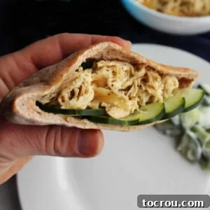 Hand holding a pita pocket filled with shredded Greek chicken and slices of cucumber.