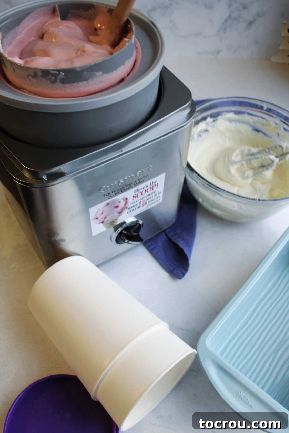 Pink Velvet Cheesecake Swirl Ice Cream 3 Ice cream maker with churned pink velvet ice cream next to mixing bowl with cream cheese frosting and some ice cream containers nearby, ready to assemble finished ice cream.