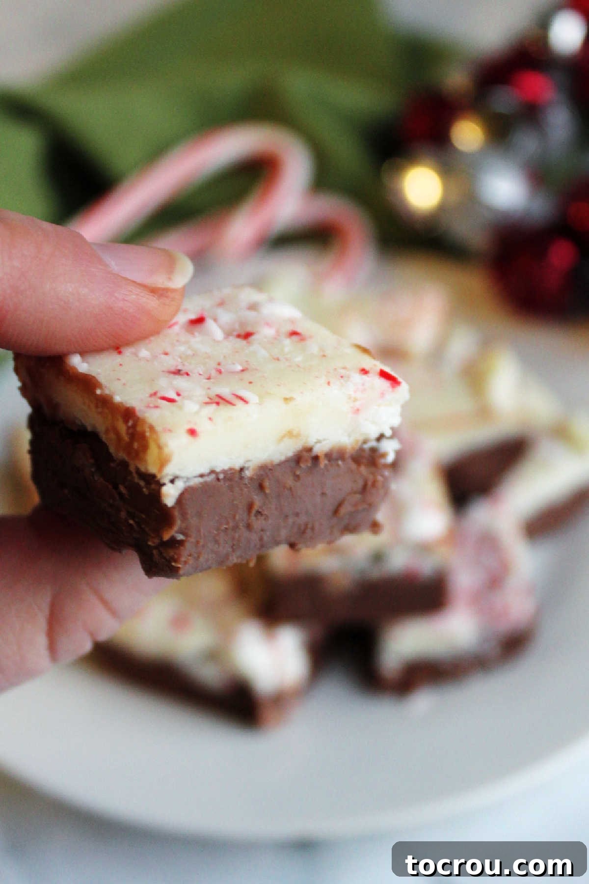 A hand holding a piece of peppermint bark fudge, showcasing its distinct layers of rich chocolate and creamy white chocolate, topped with crushed candy cane pieces.