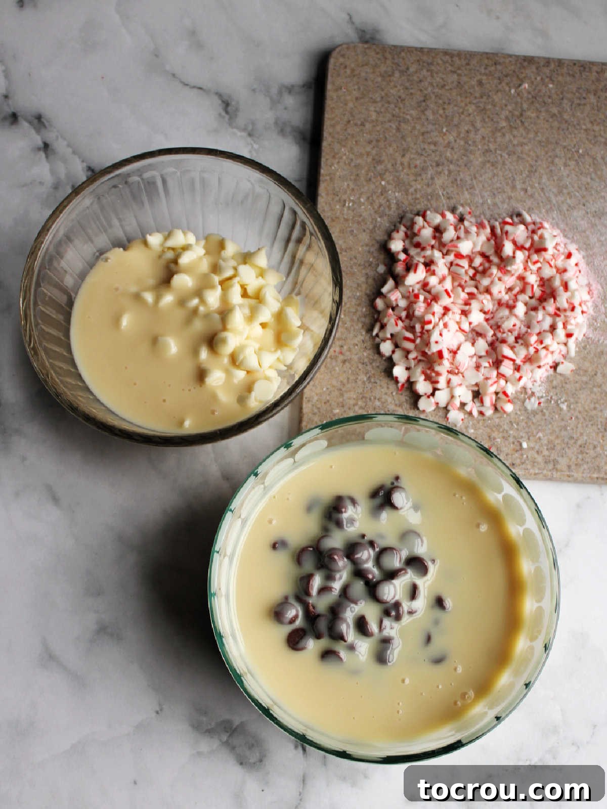 Sweetened condensed milk divided into two bowls, alongside chocolate chips, white chocolate chips, and crushed candy canes on a cutting board, ready for fudge preparation.