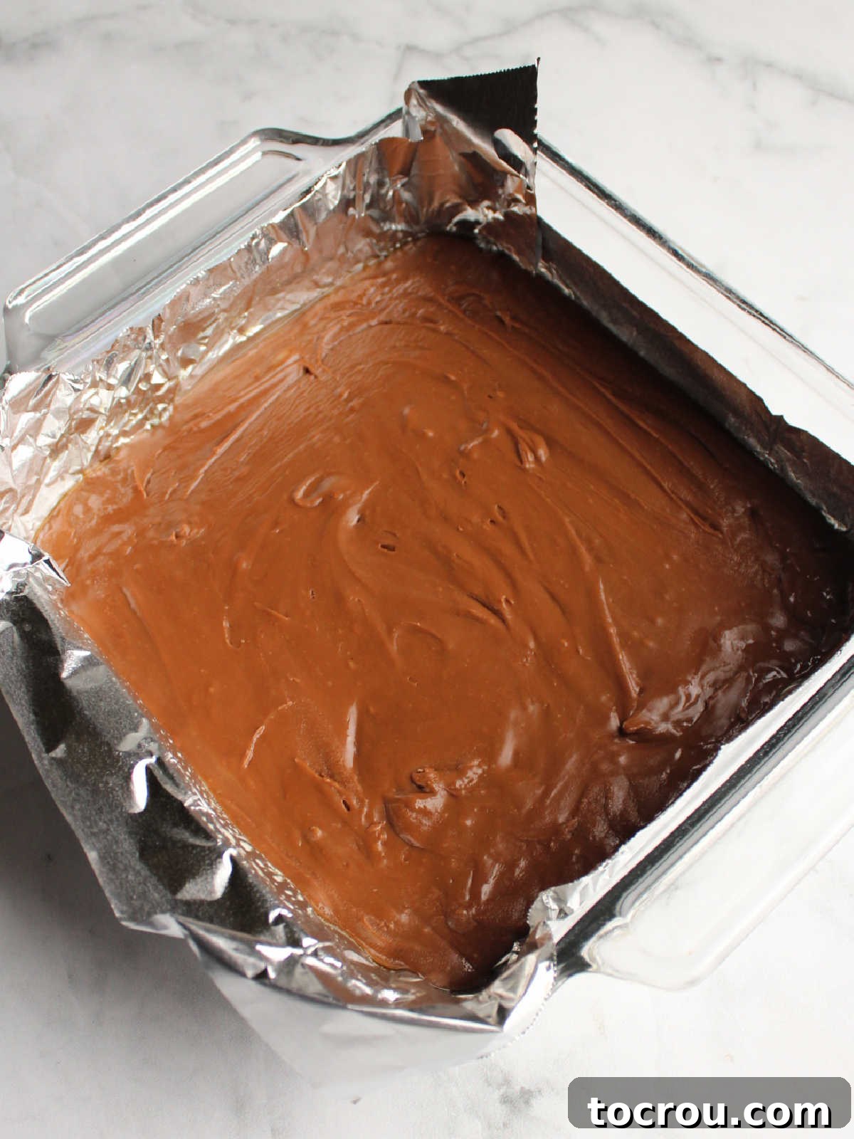 A smooth, even layer of chocolate fudge spread into an aluminum foil-lined baking pan, forming the base for the peppermint bark fudge.