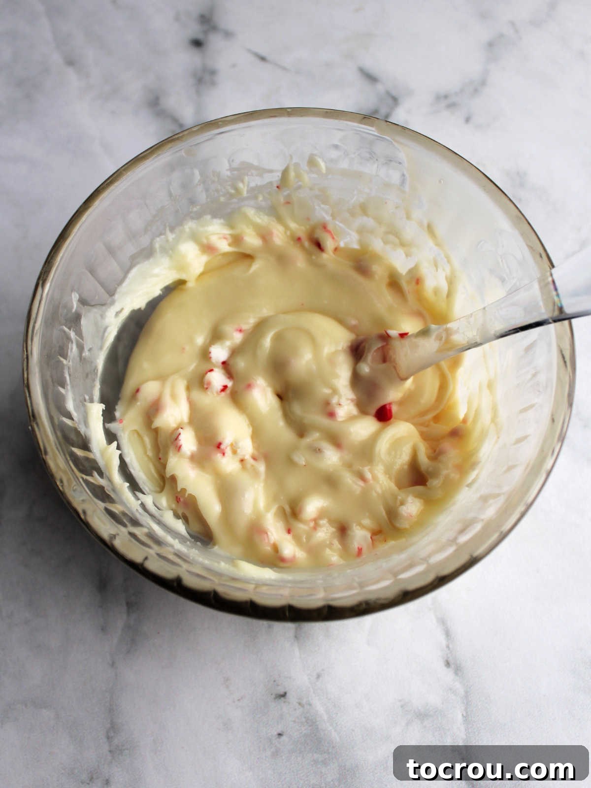 A bowl of warm, creamy white chocolate fudge with bright red and white candy cane bits stirred throughout, ready to be spread.