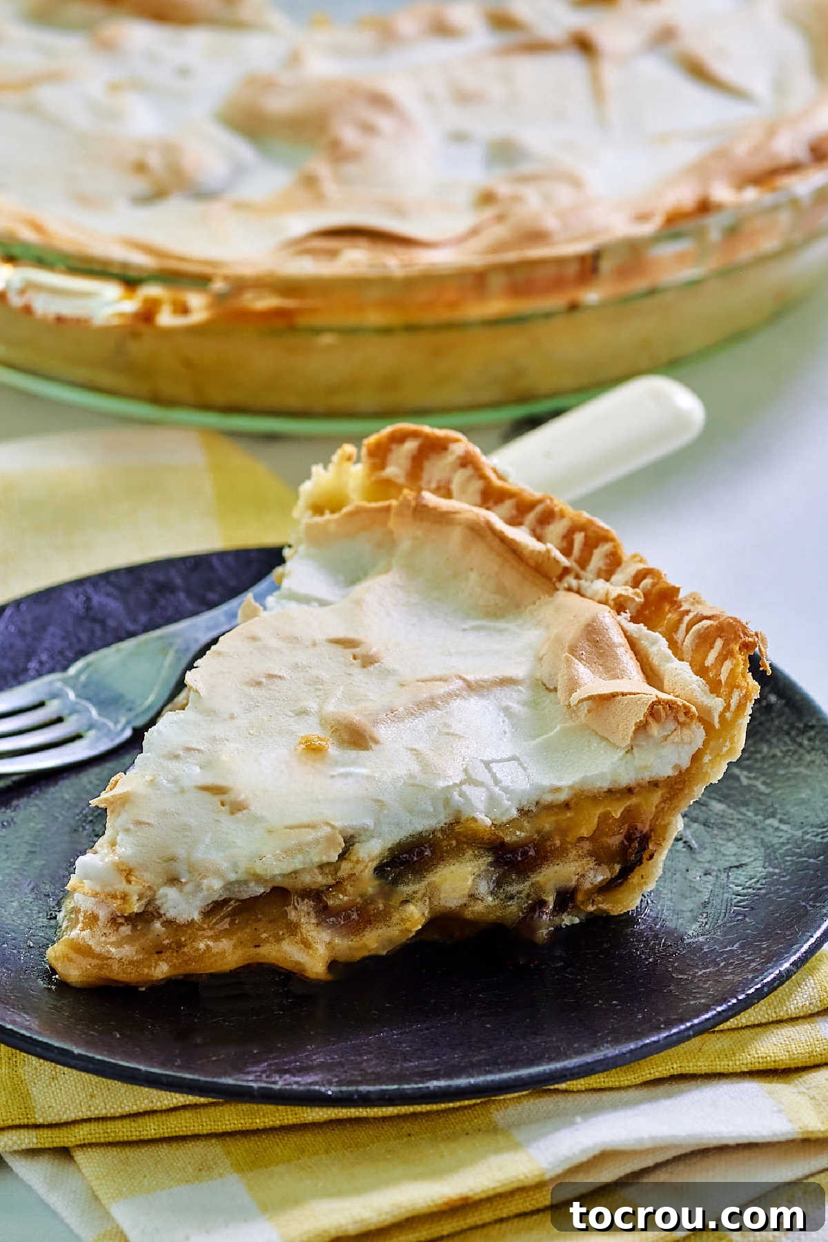 A full sour cream and raisin pie with a generous meringue topping, alongside a single slice on a dessert plate with a fork, tempting to be eaten.