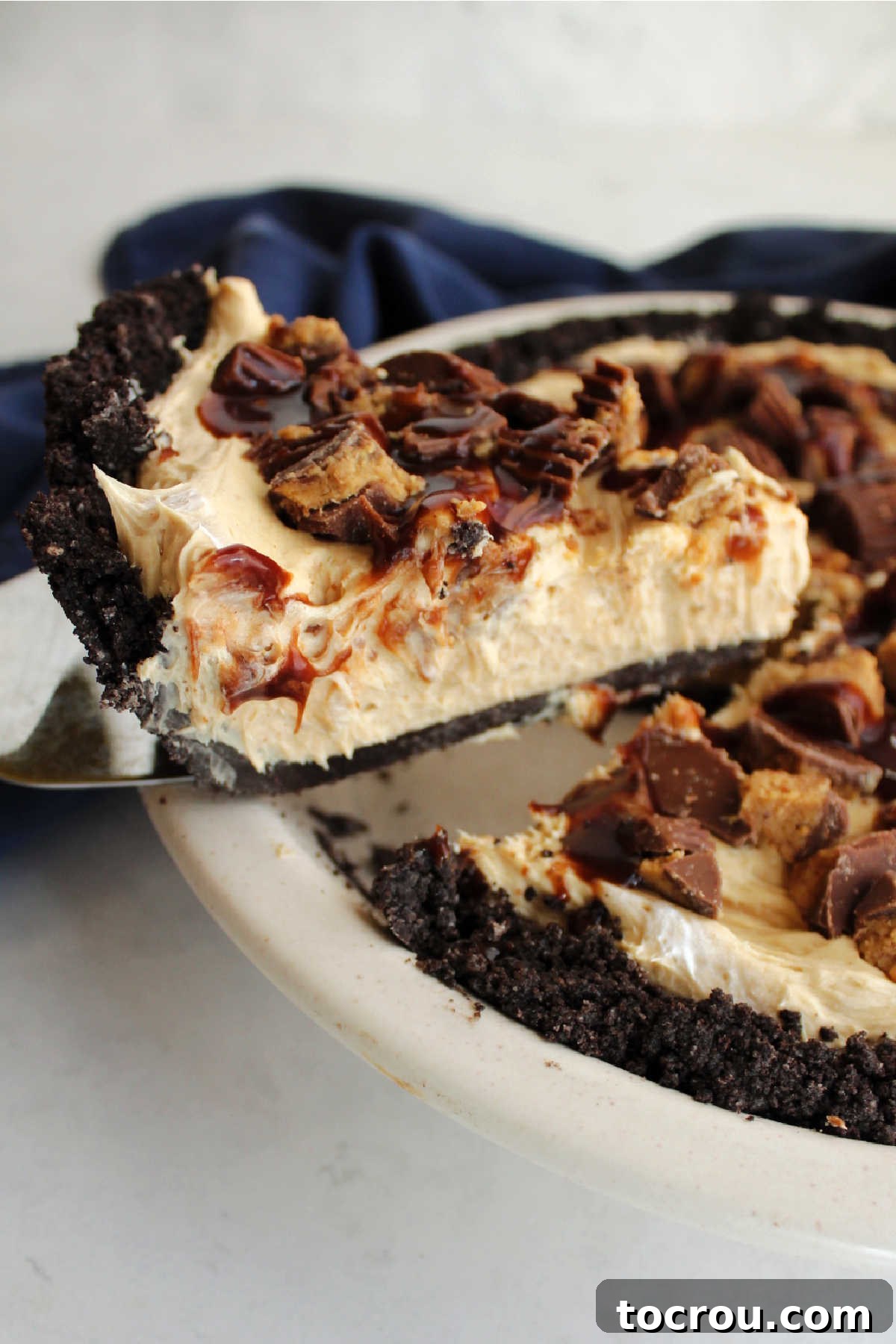 Lifting a slice of no-bake peanut butter pie from the pan, showcasing the Oreo crust, creamy peanut butter filling, and toppings of peanut butter cups and hot fudge.