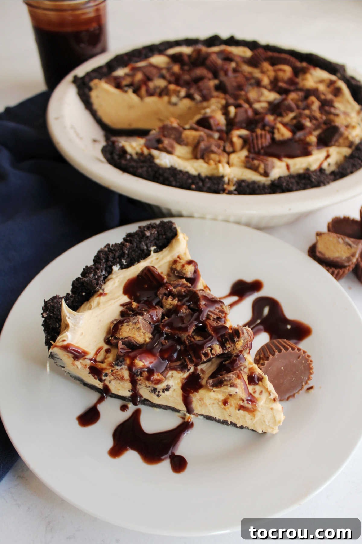 Close-up of a slice of no-bake peanut butter pie, topped with chopped peanut butter cups and drizzled with hot fudge sauce.
