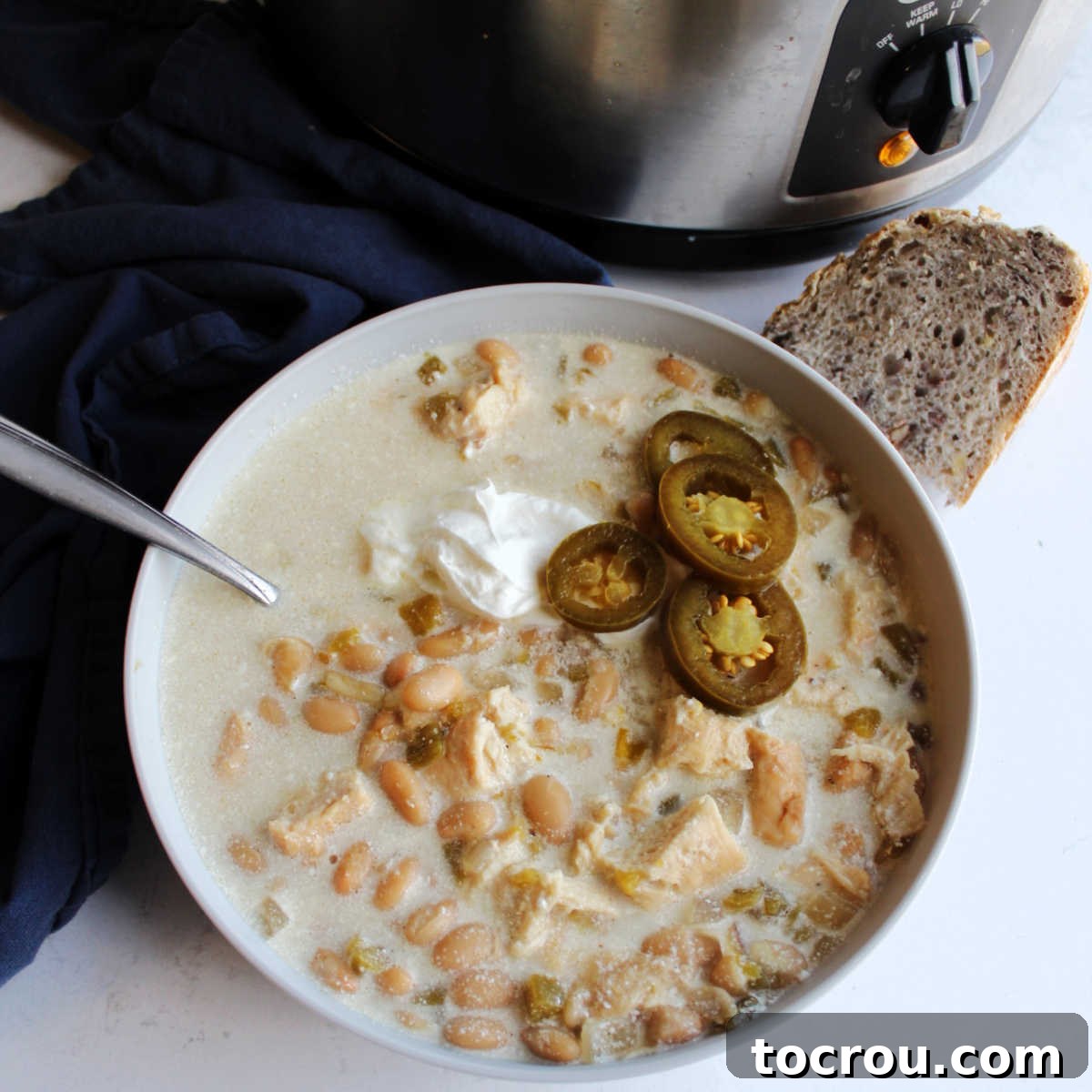 Crockpot White Chicken Chili Bowl of creamy slow cooker white chicken chili served with a dollop of sour cream, vibrant jalapeno slices, and a piece of crusty sourdough bread, all placed in front of the slow cooker it was prepared in.
