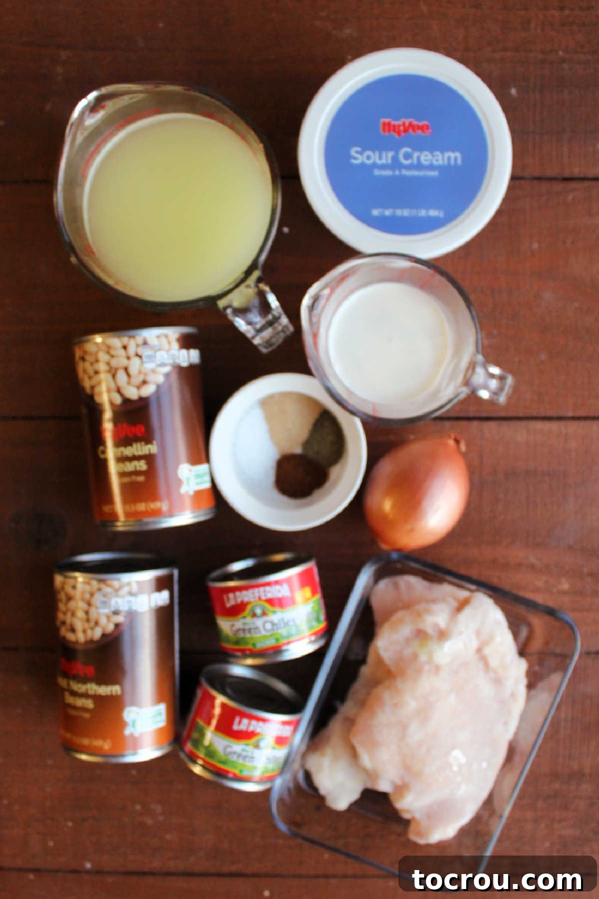 White Chicken Chili Ingredients Various ingredients for white chicken chili laid out: raw chicken breasts, canned green chilies, canned white beans, a diced onion, a selection of spices, chicken broth, and a tub of sour cream.