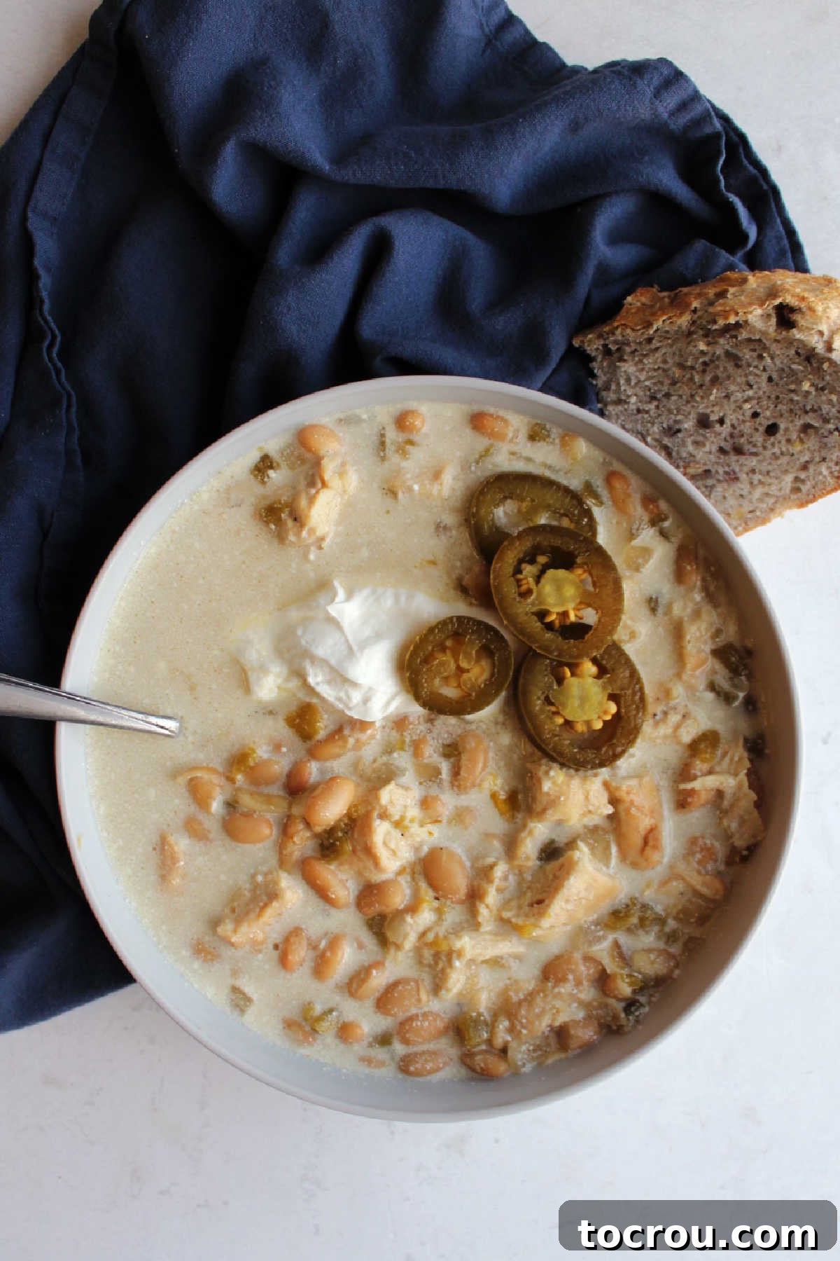White Chicken Chili with Toppings Close-up of a bowl of creamy slow cooker white chicken chili, generously topped with a dollop of sour cream and several vibrant green pickled jalapeno slices, ready to be enjoyed.